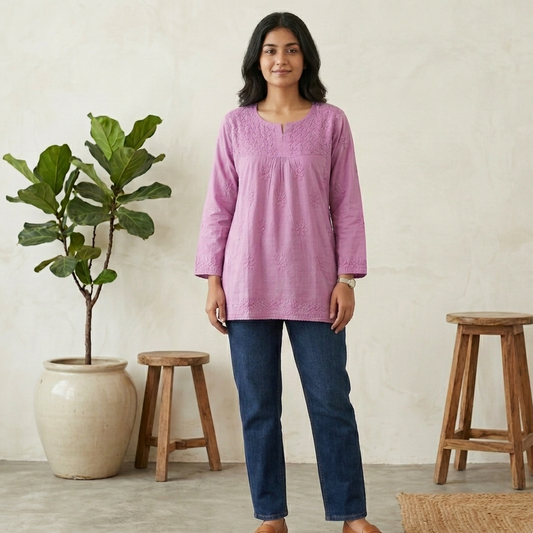 Woman wearing a purple short chikan kurti and blue jeans standing in a room with wooden stools and a plant.