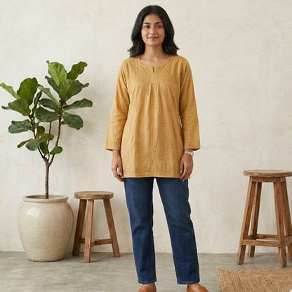 Woman wearing a GOLDEN short chikan kurti and blue jeans standing in a room with wooden stools and a plant.